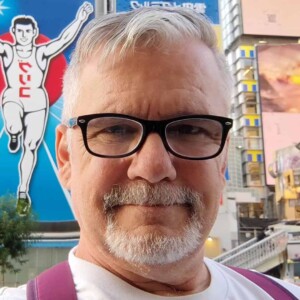 A man with glasses and a beard stands in Shibuya, Tokyo, in front of colorful signs and bright billboards, highlighting the vibrant charm of the bustling area.