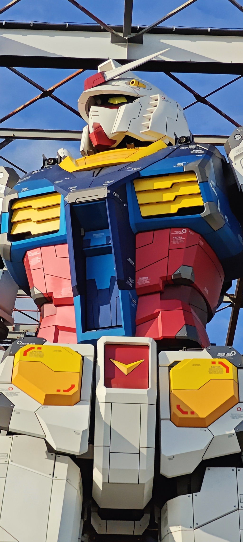 A close-up, low-angle view of a large, colorful Gundam robot statue in Tokyo, Japan, featuring white, yellow, red, and blue armor panels against a blue sky with a metal framework overhead&mdash;a true Solo Sojourn marvel.