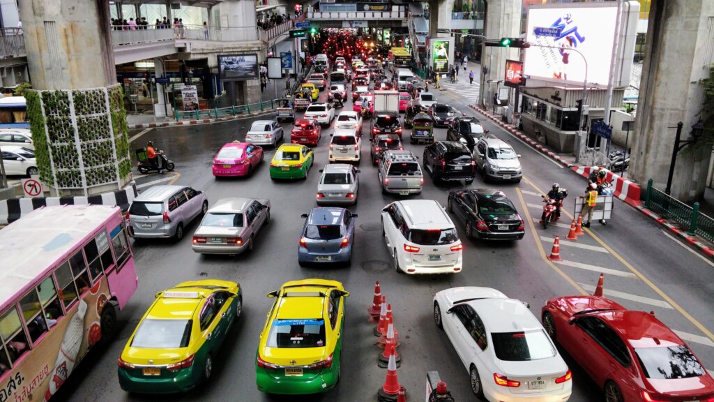 Heavy traffic with cars, taxis, and motorcycles fills a city street under an overpass in Thailands Heartbeat. Bright billboards, orange traffic cones, and pedestrians define this vibrant urban setting.